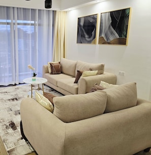 Ein modernes Wohnzimmer mit einem beigefarbenen Sofagarnitur, einem kleinen runden Couchtisch und einem weißen Teppich. An der Wand hängen zwei abstrakte Gemälde und ein Fenster mit durchscheinenden Vorhängen.