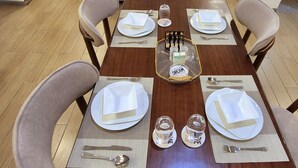 Zona pranzo con tavolo in legno apparecchiato per quattro, circondato da sedie. Sullo sfondo, un divano e una televisione.