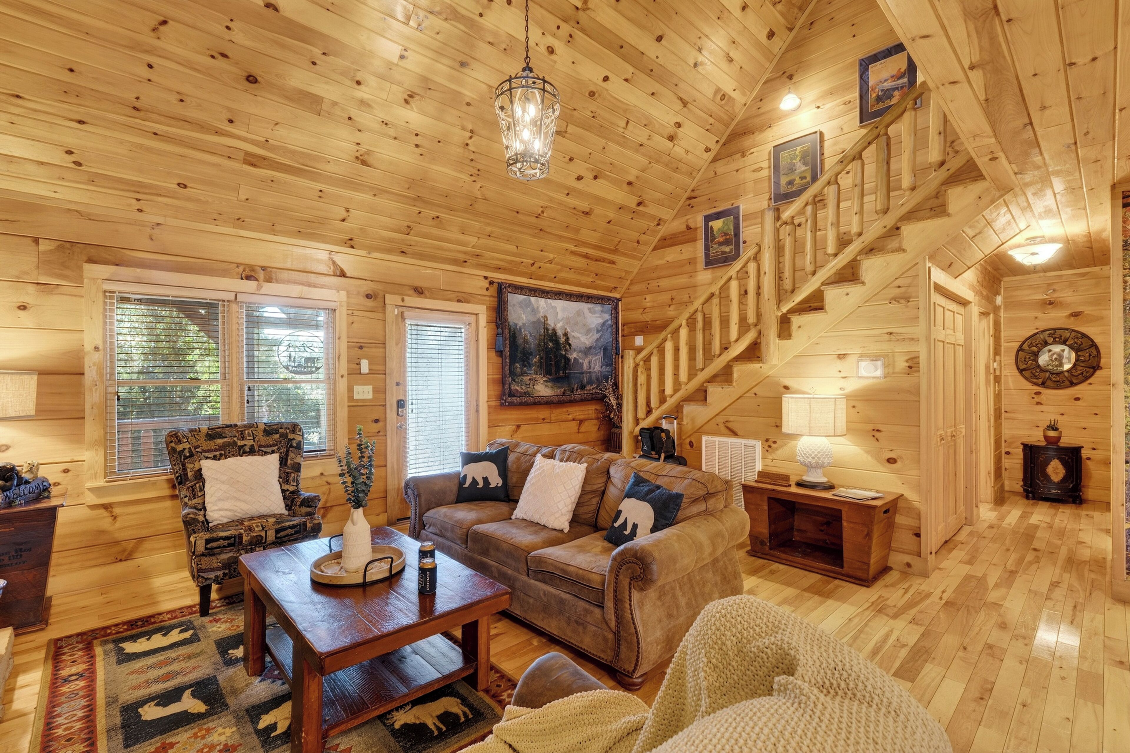 A cozy log cabin interior with a wooden staircase, a plush sofa, a patterned armchair, and a dining table.