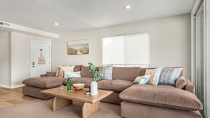 A living room with a brown sectional sofa, a wooden coffee table, and a window with blinds. - Top-Floor West Newport Retreat | Balcony | Near Beach + Cafes | Seabright Sky (Newport Beach)