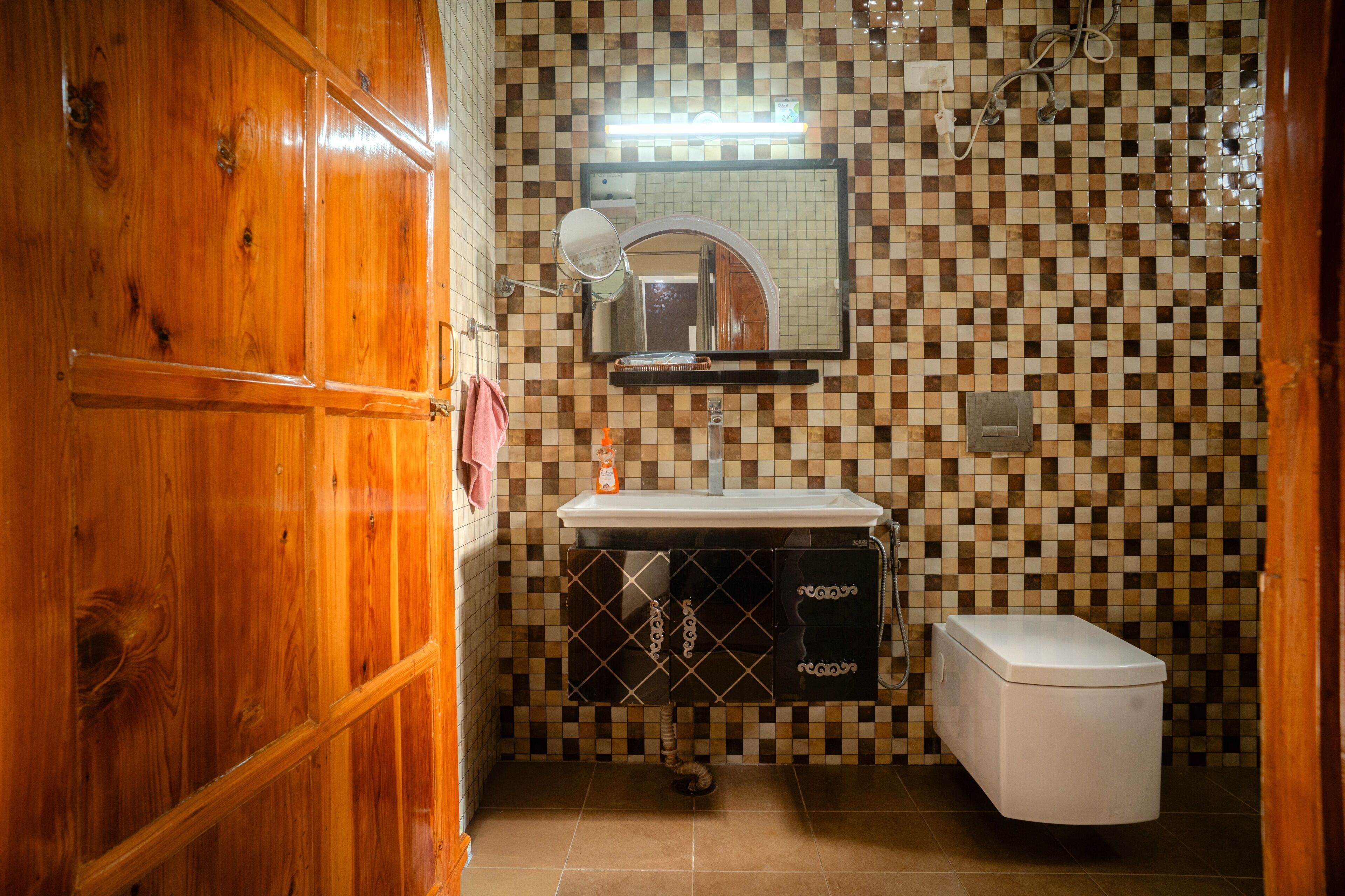 Un baño con puerta de madera, pared con azulejos, lavamanos, espejo y retrete.