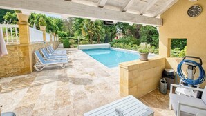Zona de piscina exterior techada con sillas de descanso, una mesa y una manguera para limpieza.