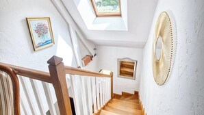 Una escalera con escalones de madera y barandillas blancas, que lleva a una habitación con tragaluces y un cuadro en la pared.