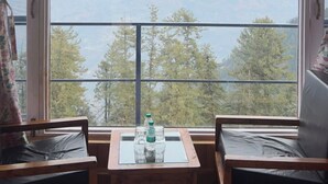 A room with a large window offering a mountain view. There is a wooden table with two chairs and a small table with bottles and glasses. - Mashobra Heights Hotel by Shalidrishya (Shimla)