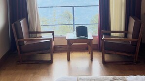 A hotel room with a large bed, two wooden chairs, a small table, and a view of mountains through a large window with curtains. - Mashobra Heights Hotel by Shalidrishya (Shimla)