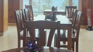 A wooden dining area with a table and chairs, a view of mountains through a large window, and a small decorative pine tree on the table. - Mashobra Heights Hotel by Shalidrishya (Shimla)