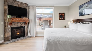 A bedroom with a stone fireplace, a large bed, a flat-screen TV, and a view of mountains through the window. - Ski-In Ski-Out Home + Hot Tub + Resort Amenities + Alpine Views | Highline Lodge (Park City)