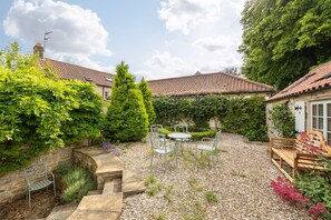 A courtyard with a stone bench, a table with chairs, and a wooden bench with cushions.