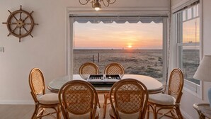 A dining area with a view of the sunset over the beach, wicker chairs, a ship's wheel, and a chandelier.