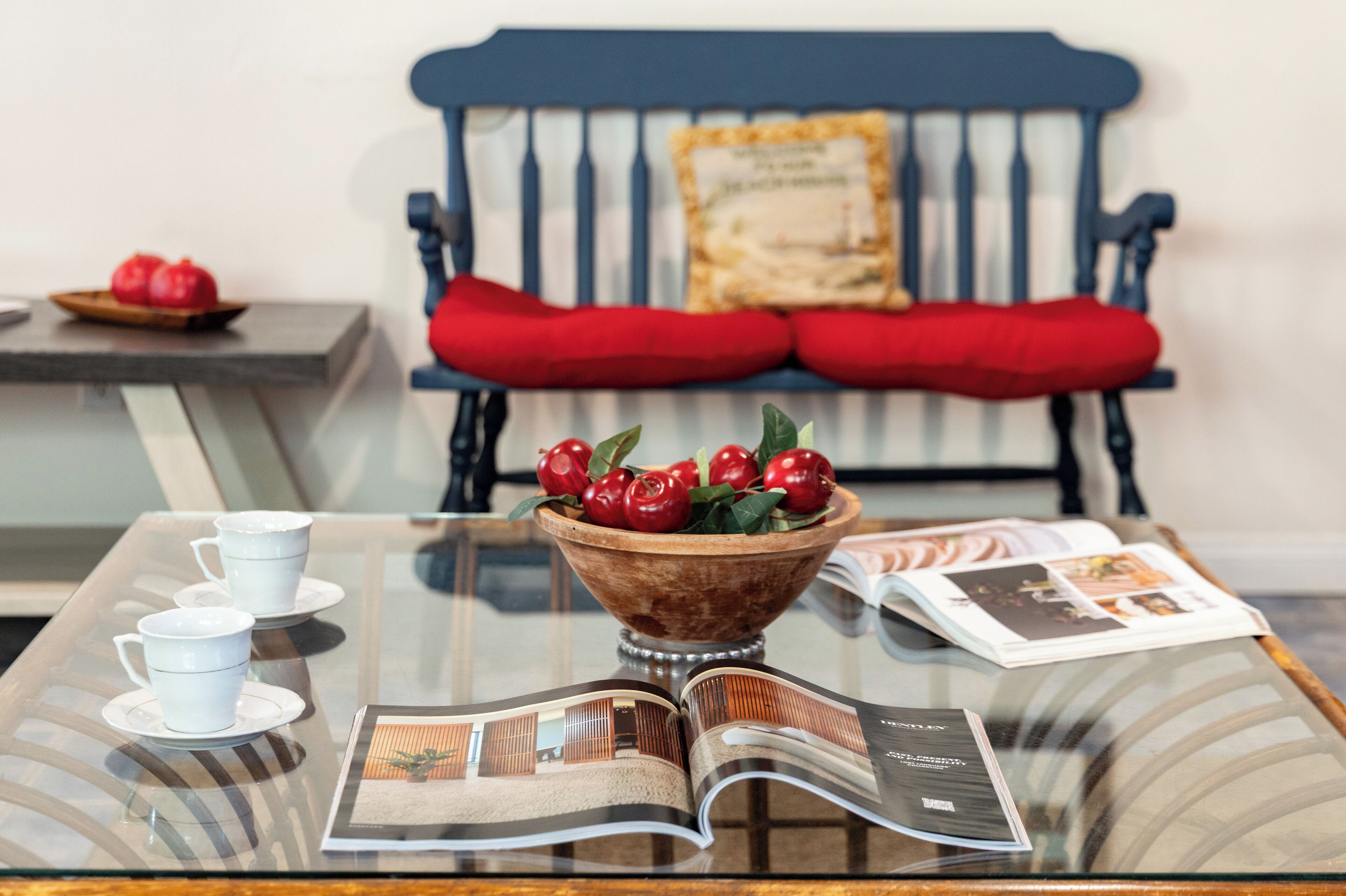 A glass-top table with a bowl of cherries, a magazine, and two white cups on saucers.