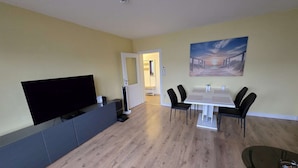 Een moderne woonkamer met een flatscreen televisie, een grijze tv-meubel en een eethoek met een witte tafel en zwarte stoelen.