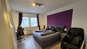 Een moderne slaapkamer met een bed, een massagestoel, een bureau met stoel en een raam met gordijnen.