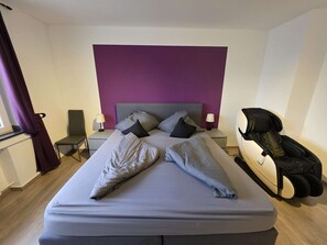 A modern hotel room with a large bed, a headboard, bedside tables, a chair, a massage chair, and a window with curtains. - Business apartment, 30+ nights – 80 m², 2 bedrooms (Leverkusen)