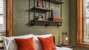 A bed with white and orange pillows, a bedside lamp, and a shelf with books and decorative items against a textured green wall. - LAST MIN DEAL - Historic Condo - 1 King 2 Queens (Greensboro)