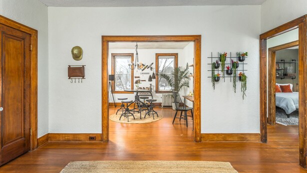 A spacious living room with hardwood floors, a central seating area with a coffee table, and a dining table. There are multiple hanging plants on the wall, a large window providing natural light, and a door leading to another room. - LAST MIN DEAL - Historic Condo - 1 King 2 Queens (Greensboro)