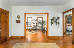 Un vaste salon doté de planchers en bois, un espace salon central avec une table basse et une table à manger. Plusieurs plantes suspendues ornent le mur, une grande fenêtre apporte une lumière naturelle et une porte donne accès à une autre pièce.