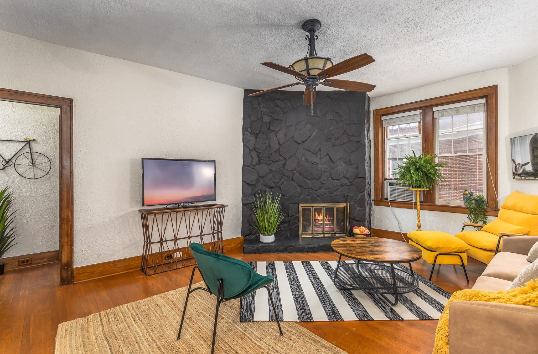 A living room with a stone fireplace, a television, a ceiling fan, and a large window.