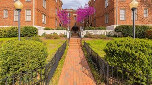 A brick building with a central pathway leading to a door, surrounded by greenery and fences. - LAST MIN DEAL - Historic Condo - 1 King 2 Queens (Greensboro)