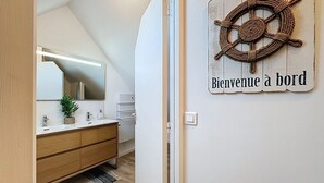 Une salle de bain moderne avec un meuble-lavabo en bois, un grand miroir et une décoration en forme de barre de navire portant l’inscription « Bienvenue à bord ».