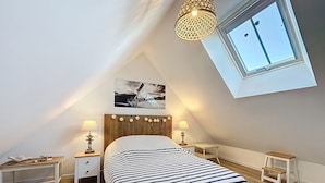 Une chambre avec un plafond en pente, un lit recouvert d’une couvre-lit à rayures, deux tables de chevet avec des lampes, un petit tabouret et une fenêtre donnant sur un immeuble.