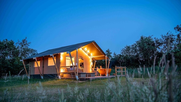 Un allotjament acollidor en estil de tenda, ben il·luminat, amb una zona de porxo, envoltat de vegetació natural.