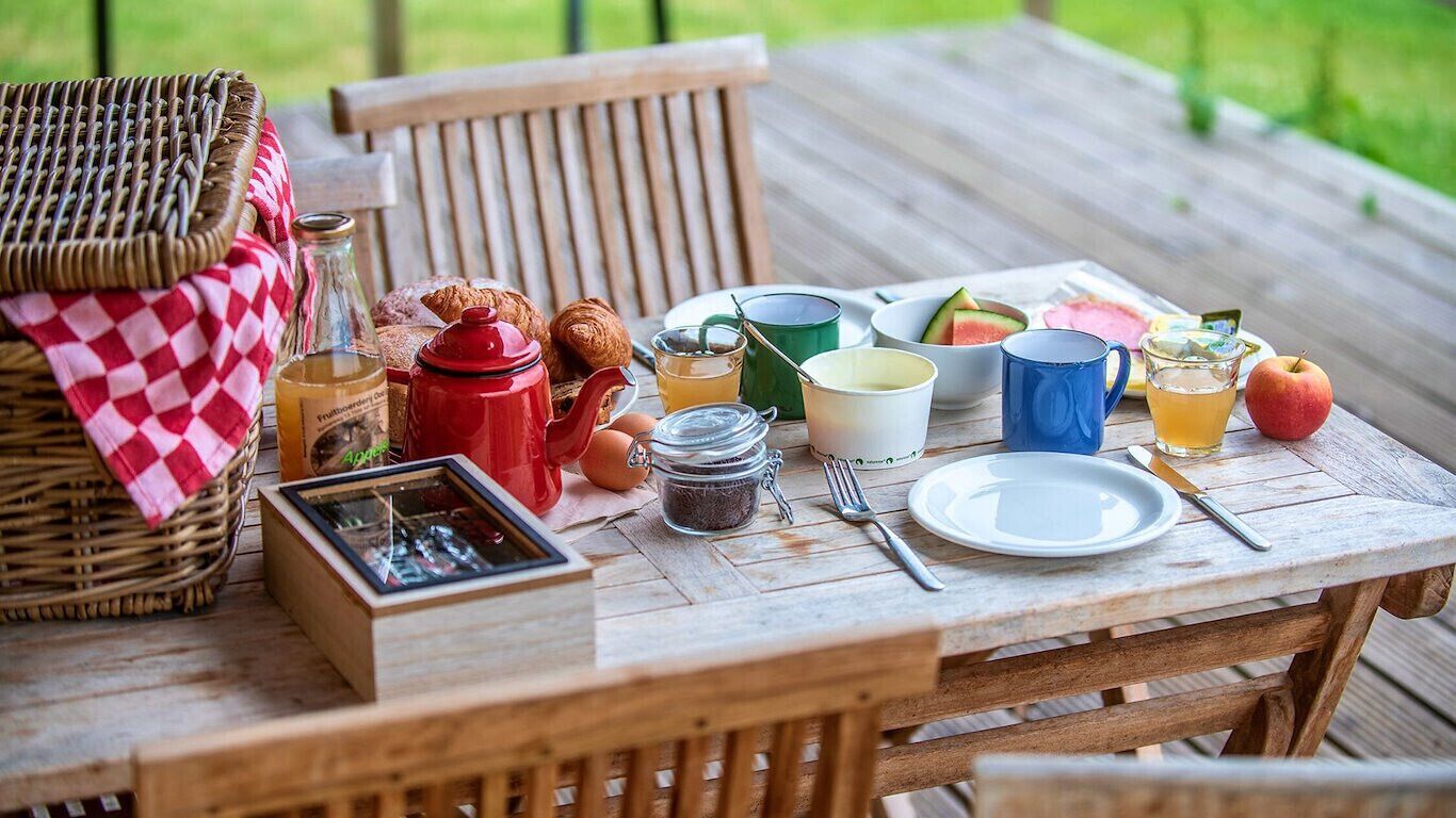 A wooden table set with breakfast items including croissants, a red teapot, a glass of juice, and a plate of food.