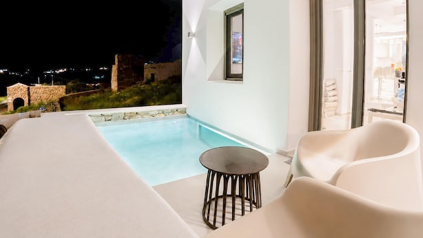 A modern outdoor pool area with a stone wall, a small round table, and two white chairs. - Eidolion Essence - Mykonos Villa with Private Pool (Mykonos)