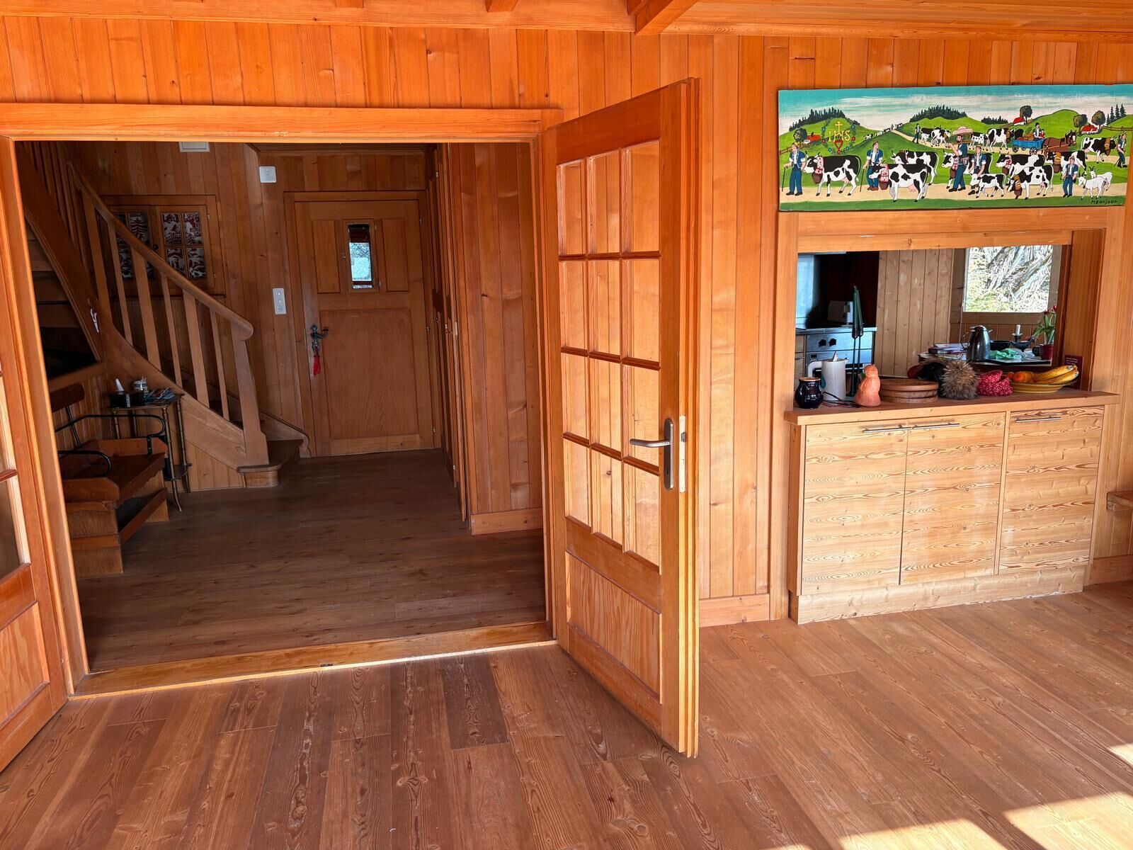 A wooden cabin interior with a wooden staircase, a kitchen area with cabinets, and a painting above the counter.