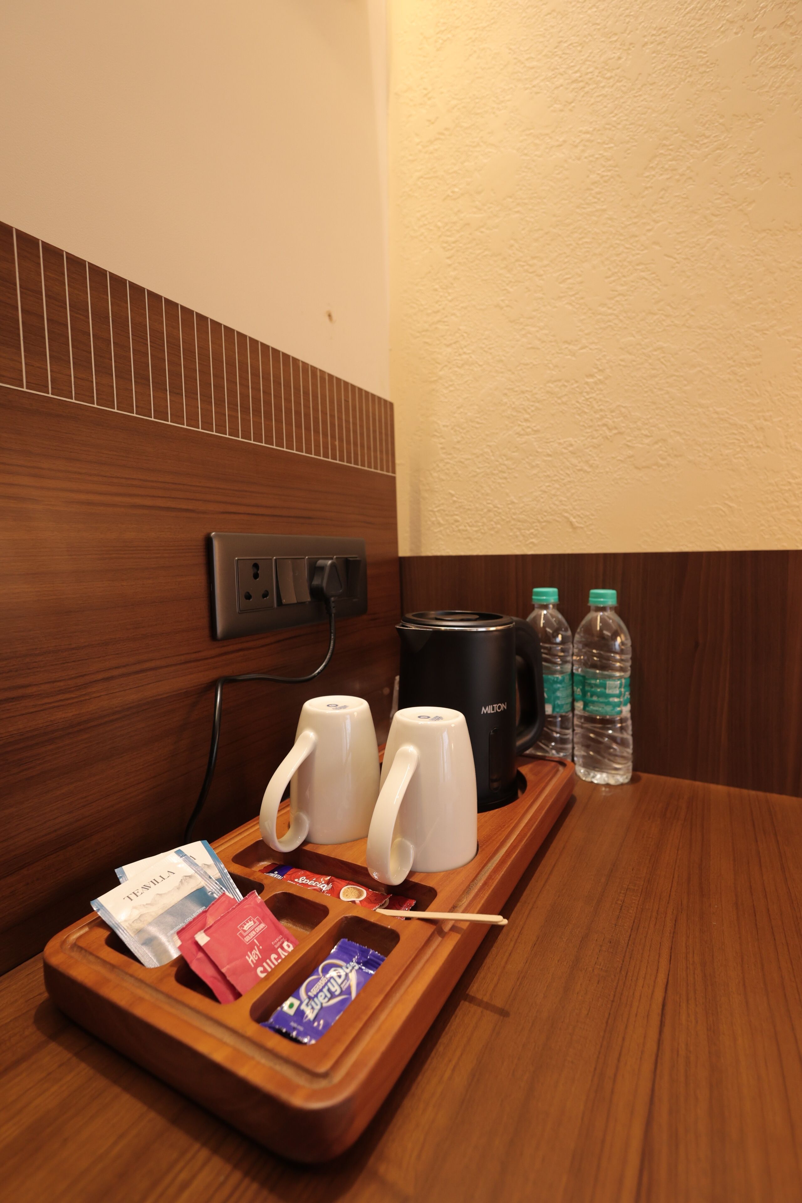 A wooden tray with a coffee maker, two mugs, a bottle of water, and a tray with snacks and a toothbrush.