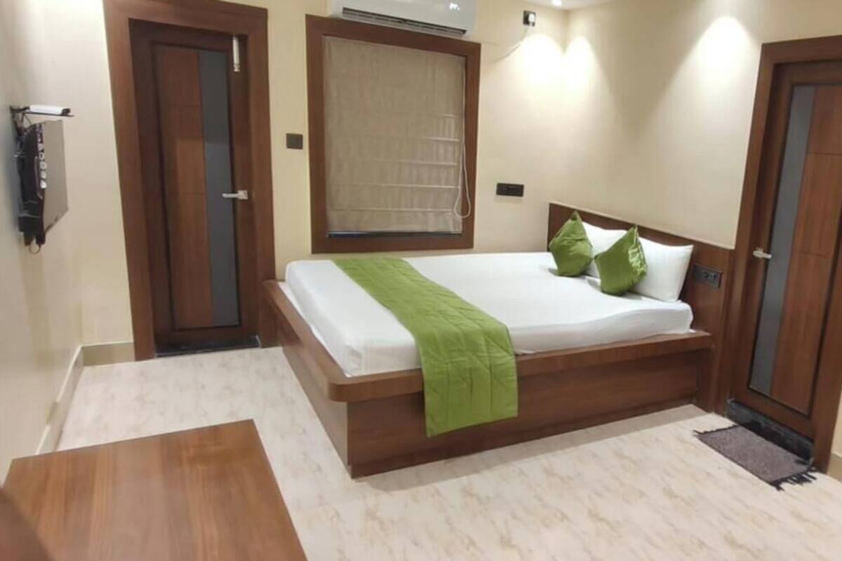 A hotel room with a bed, wooden headboard, a green throw, a window with blinds, and a door.