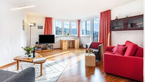 Een moderne woonkamer met een flatscreen televisie, een houten bureau, een rode bank en grote ramen met gordijnen.