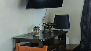 Ein Zimmer mit einem wandmontierten Flachbildfernseher, einem Tisch mit Lampe und einem Fenster mit Vorhängen.