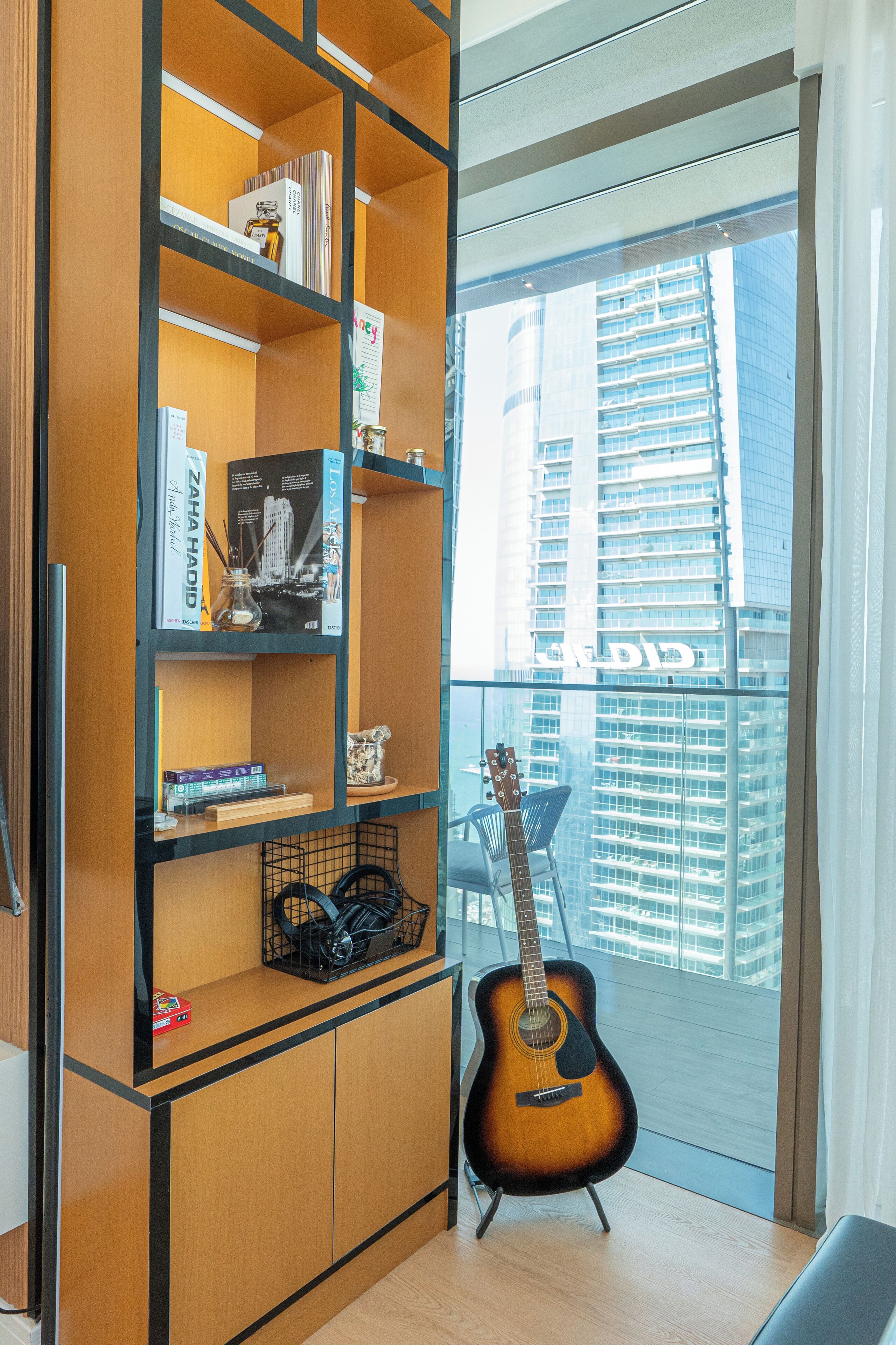A room with a wooden bookshelf, a guitar, and a view of a cityscape through a large window.