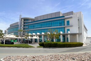 A modern building with multiple floors, large windows, and a parking area with cars and covered parking structures.