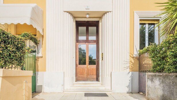 Una entrada de edificio con puerta de madera, un porche blanco y una valla verde.