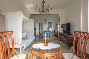 Une pièce avec un lit recouvert de draps blancs, une table à manger avec des chaises, un lustre et un téléviseur.