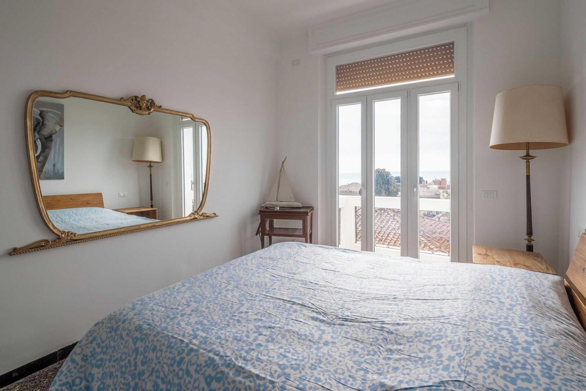 Une chambre à coucher avec un lit, un grand miroir, une fenêtre avec des stores et une lampe.