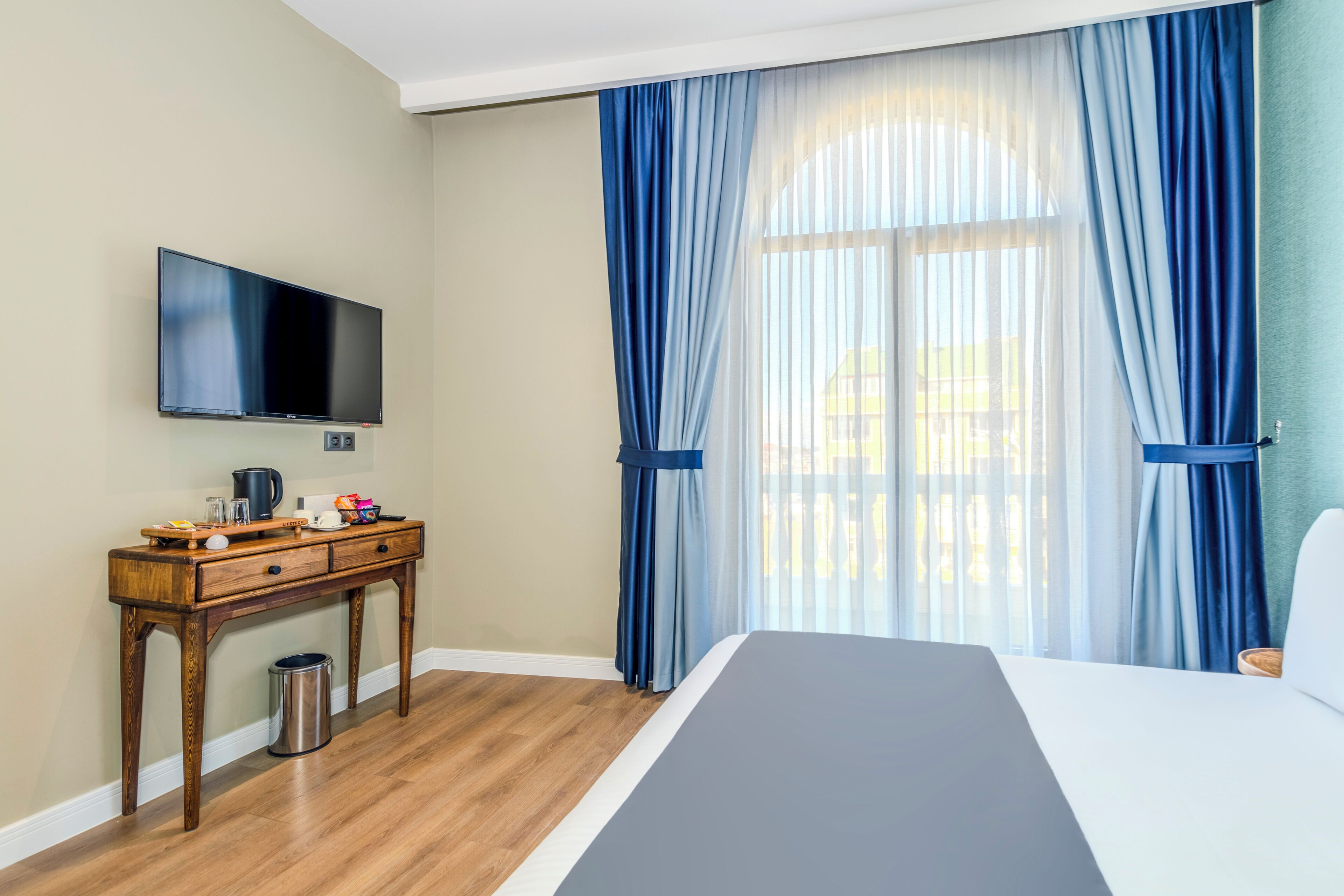 A hotel room with a bed, a wooden desk with a flat-screen TV, a trash can, and a window with blue curtains.