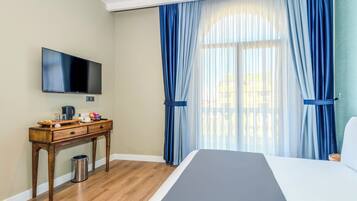 A hotel room with a bed, a wooden desk with a flat-screen TV, a trash can, and a window with blue curtains.
