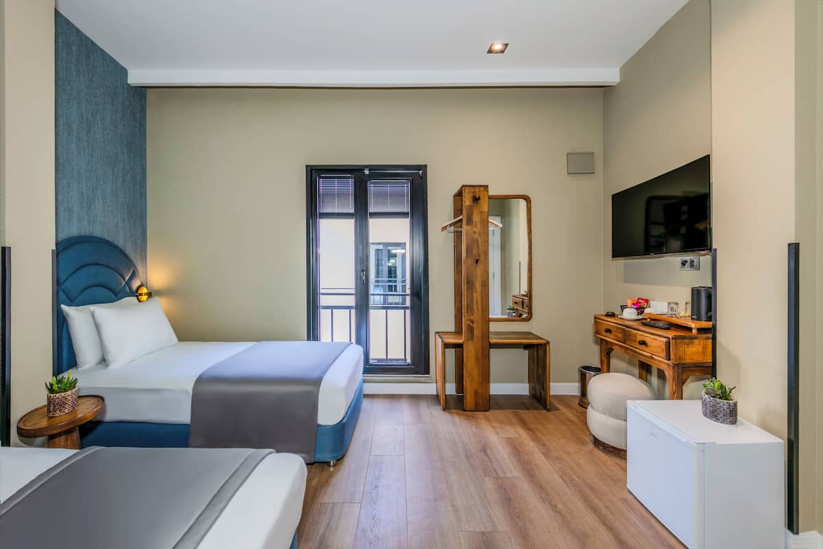 A modern hotel room with a bed, a desk, a mirror, a TV, and a window with blinds.