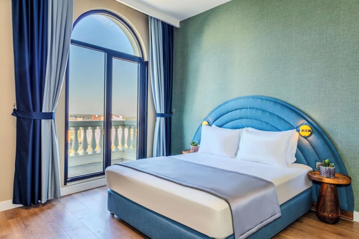A hotel room with a large bed, a nightstand, a potted plant, and a view of a balcony with a railing.