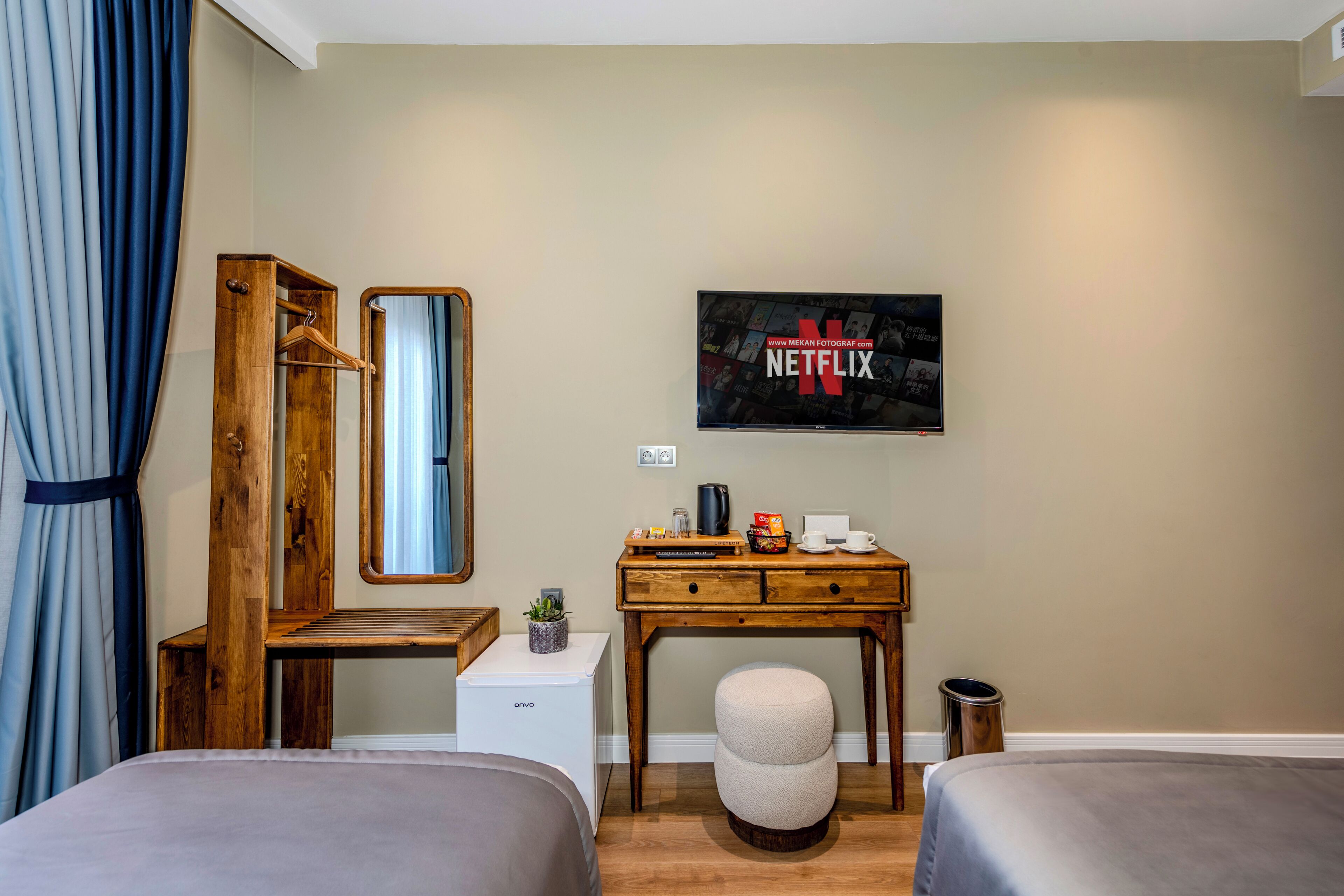 A hotel room with two beds, a wooden desk, a small refrigerator, a TV mounted on the wall, and a trash can.