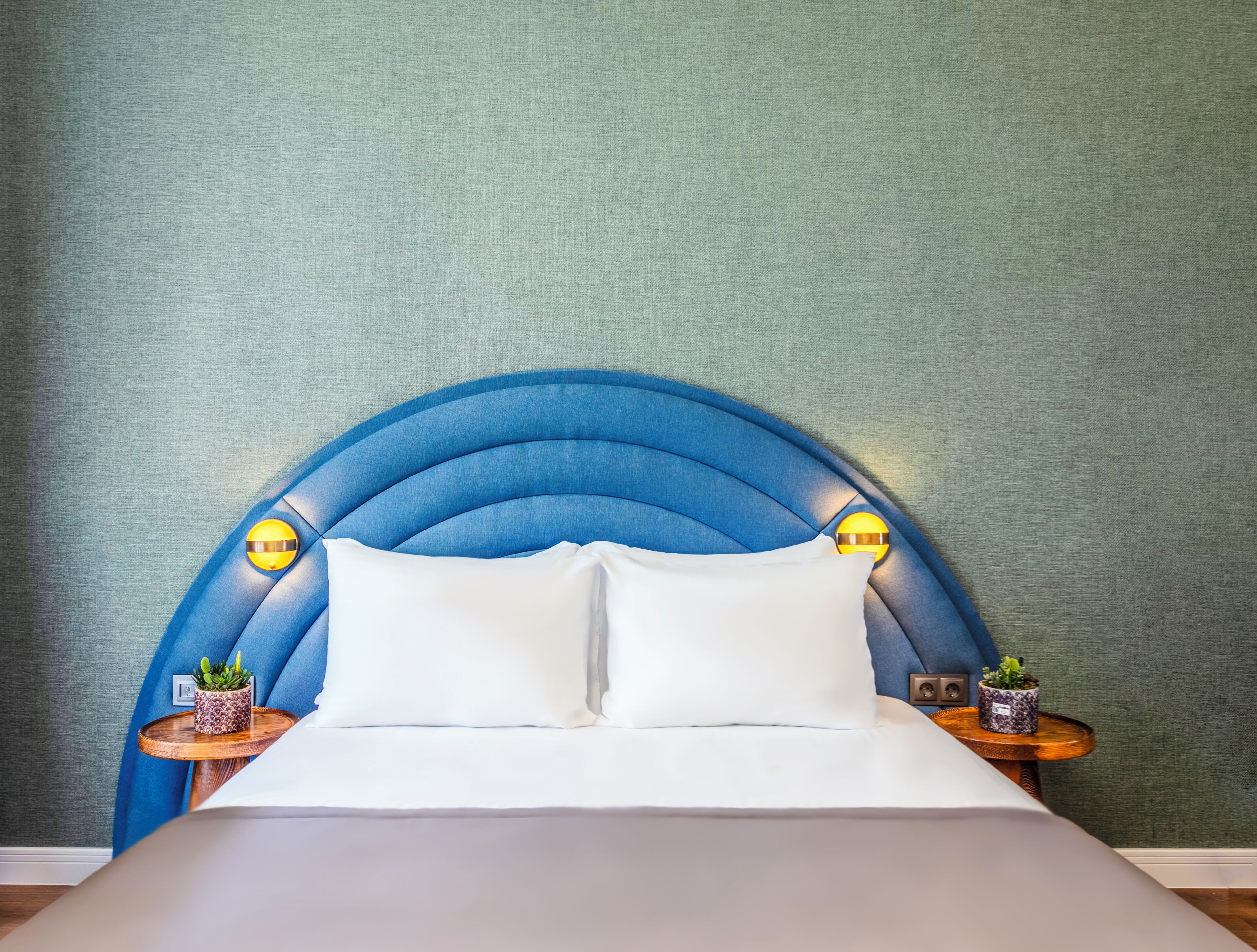 Een bed met een blauw bekleed hoofdbord, twee nachtkastjes met lampen, en een groene, gestructureerde wand.