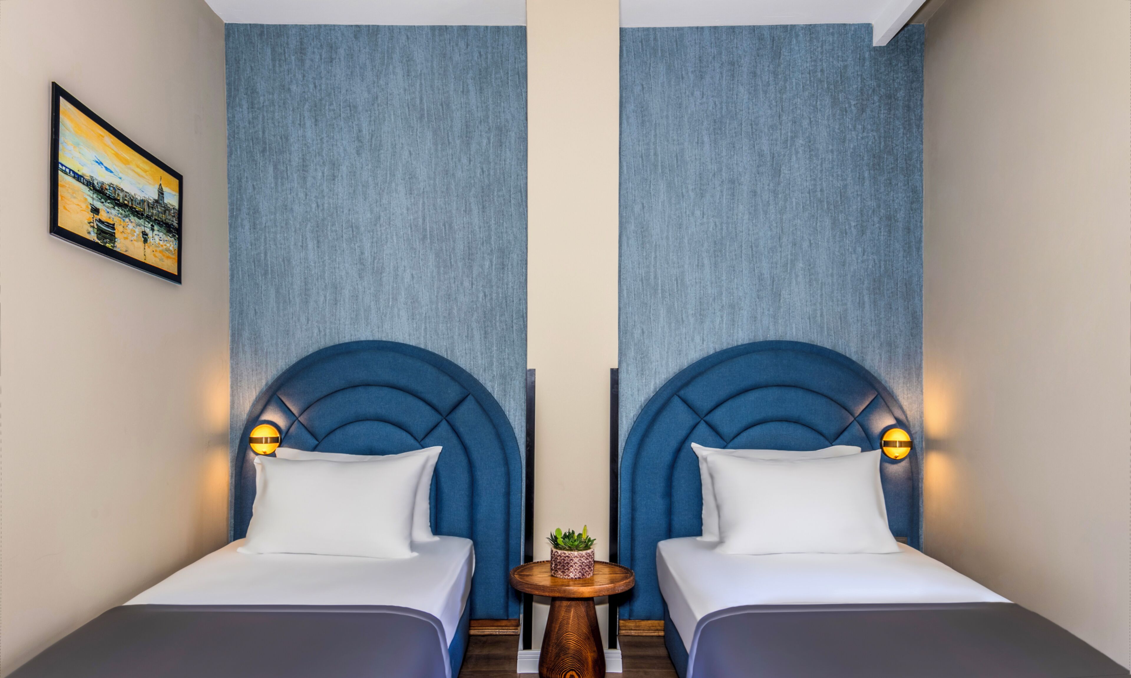 Een hotelkamer met twee bedden, elk voorzien van een blauwe hoofdbord en witte beddengoed. In het midden van de kamer staat een klein rond tafeltje met een plant.