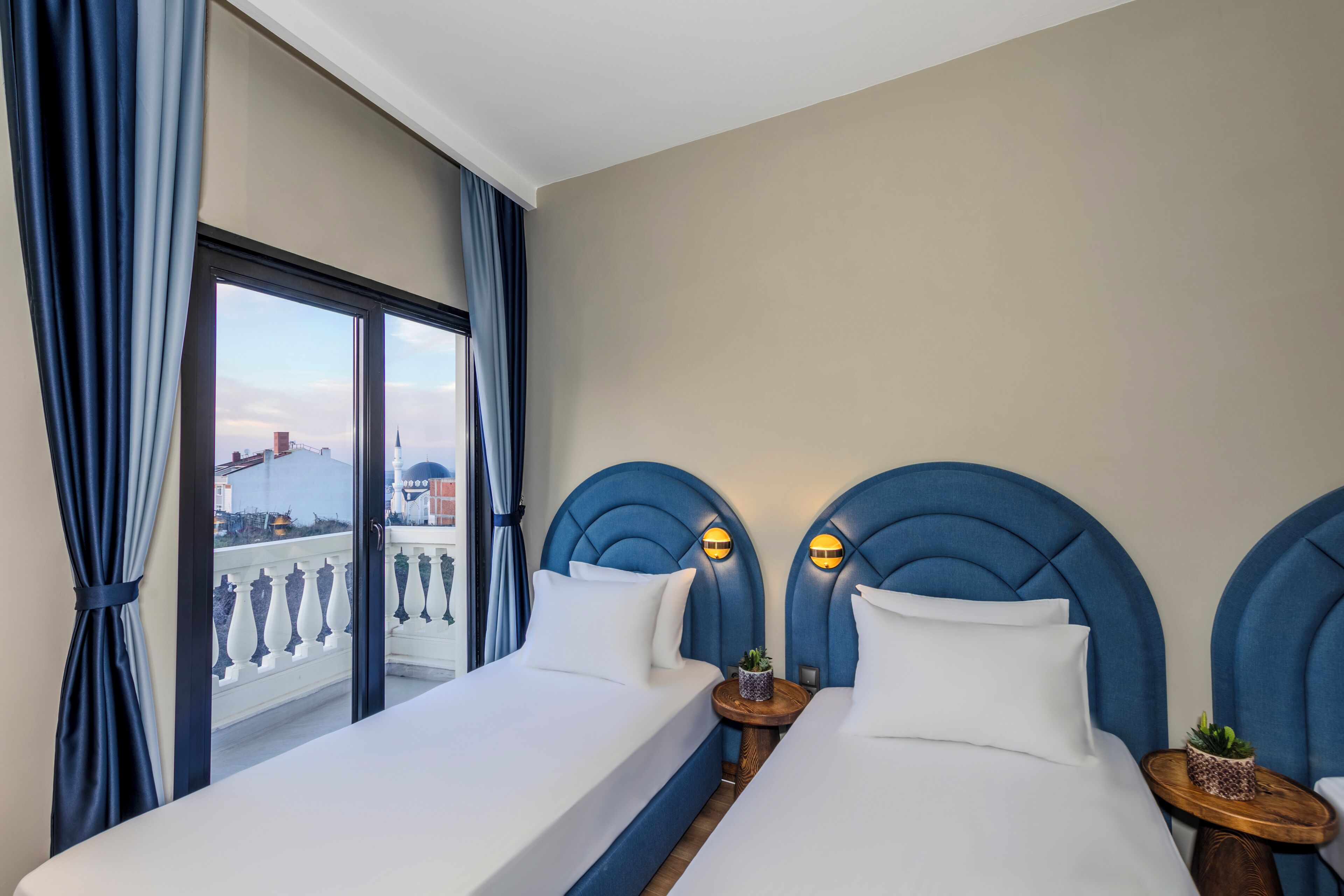 Een hotelkamer met twee bedden, een balkon met een reling en uitzicht op gebouwen.