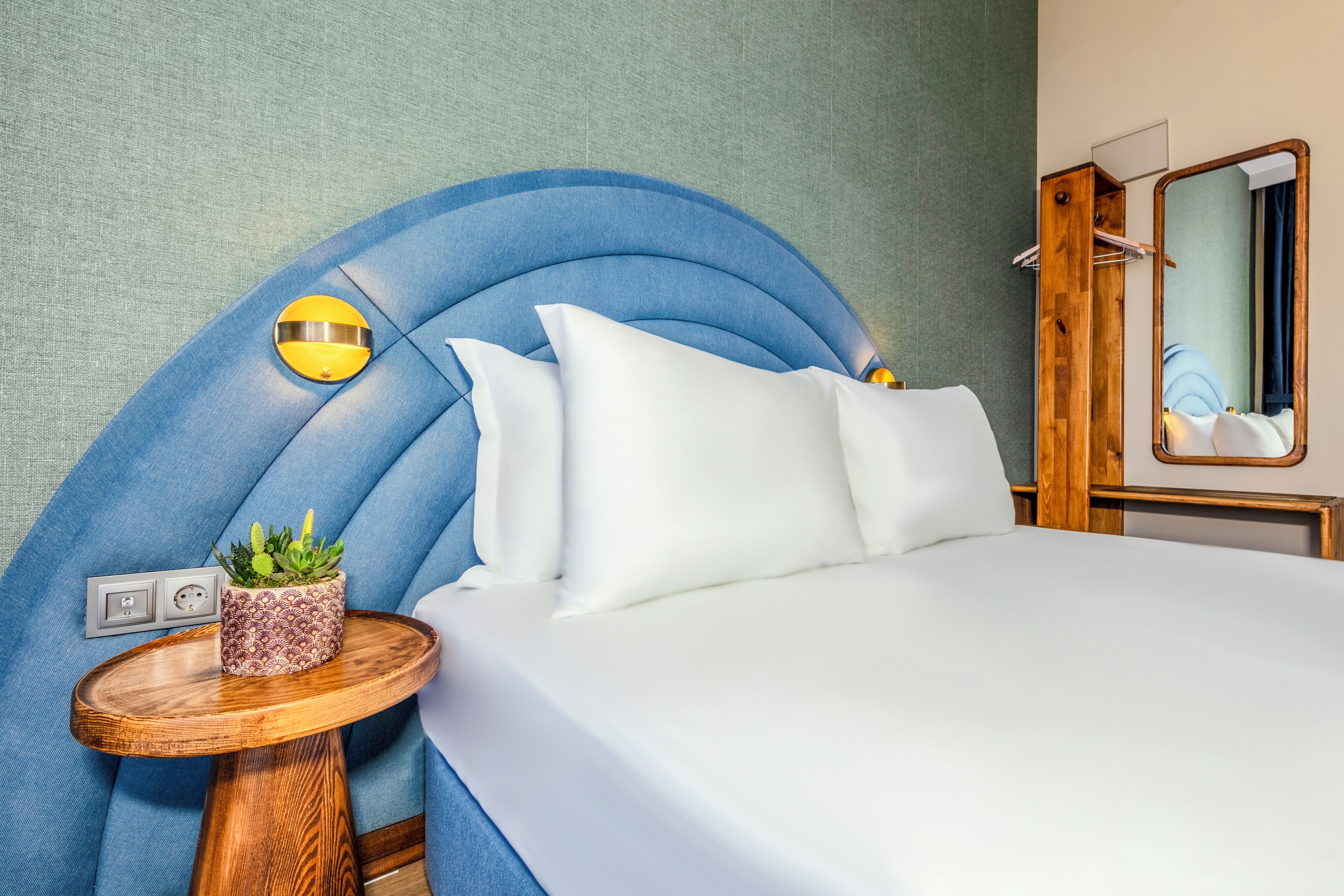 A neatly made bed with white linens, a round wooden side table with a potted plant, and a curved blue headboard.