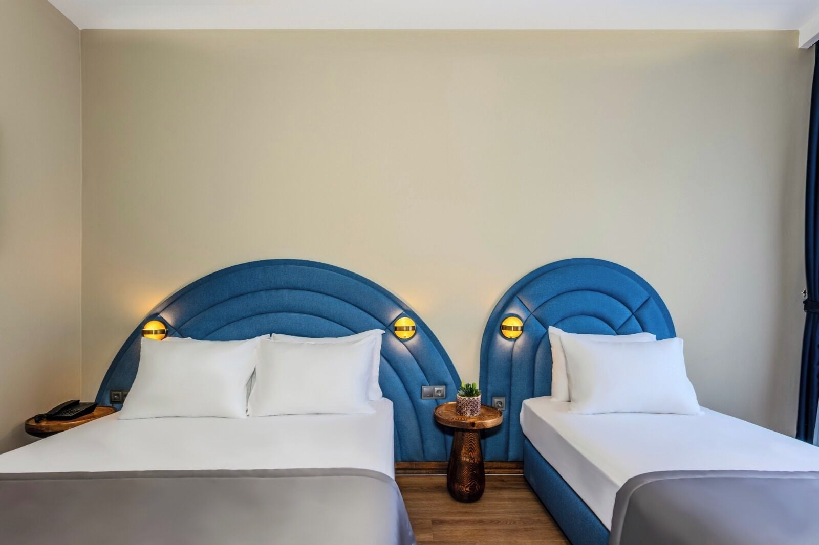 Een hotelkamer met twee bedden, elk voorzien van een blauw hoofdeinde en witte beddengoed. Tussen de bedden staat een klein houten nachtkastje met een potplant.