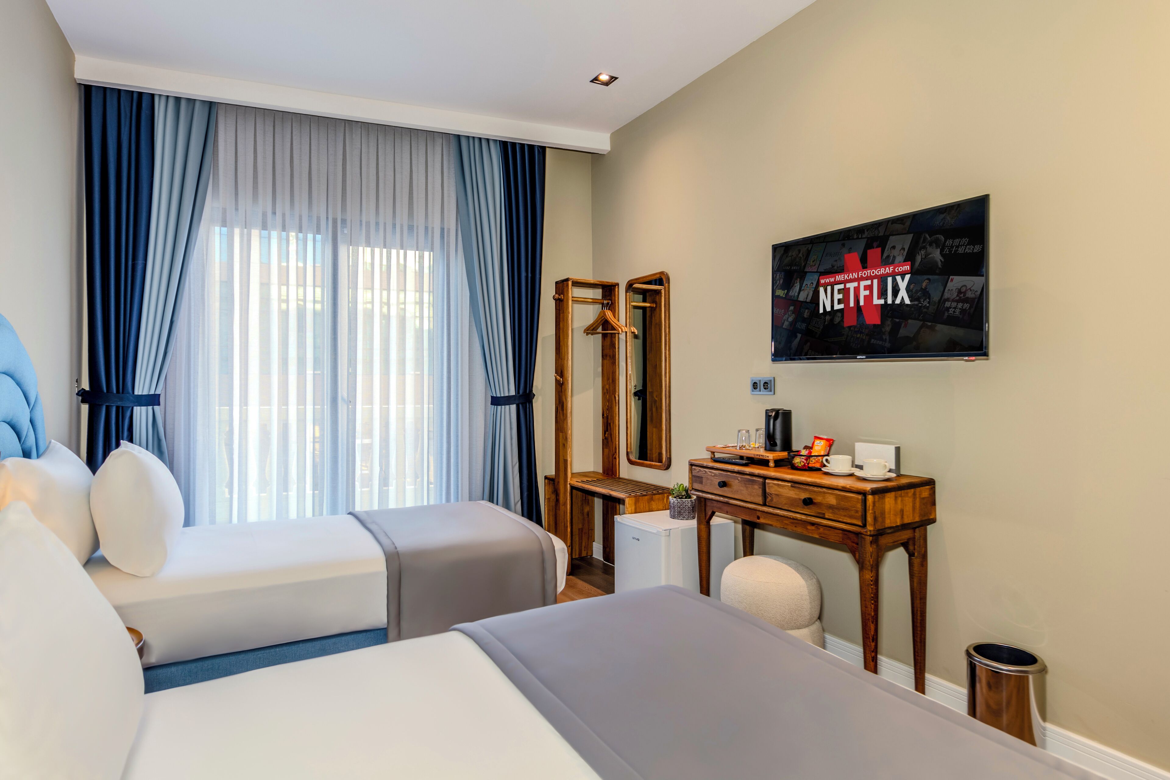 Une chambre d’hôtel avec deux lits, un bureau en bois, un miroir, un petit réfrigérateur et un téléviseur fixé au mur qui affiche le logo de Netflix.