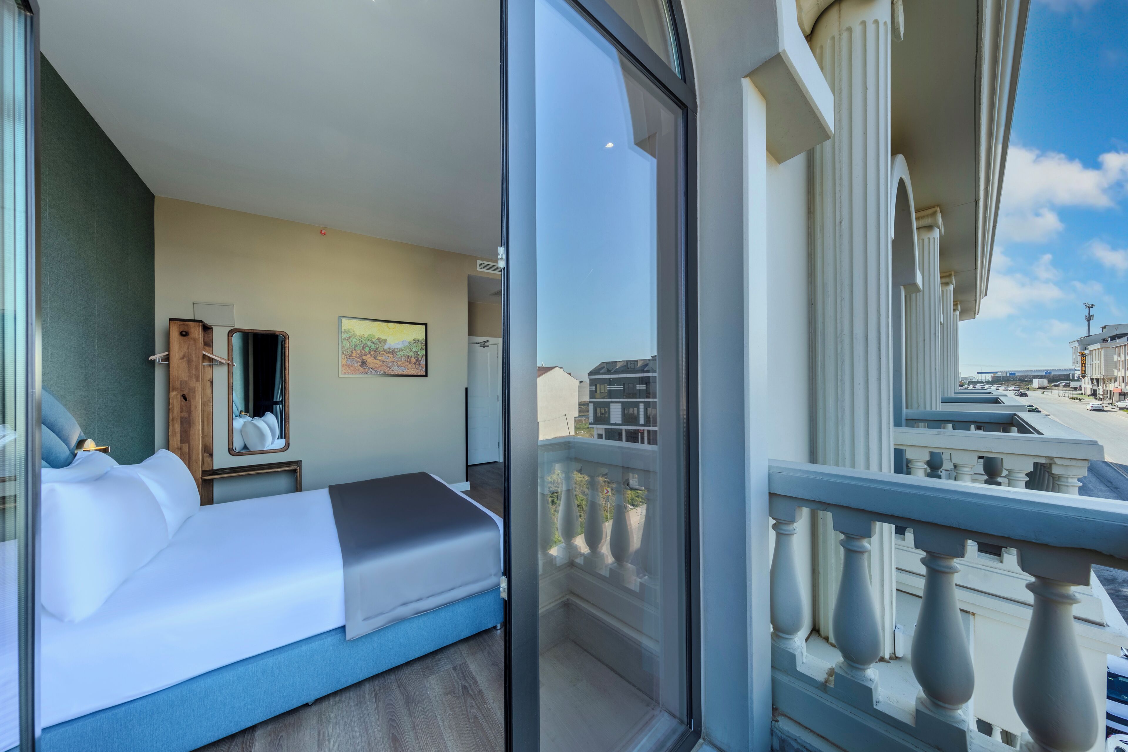 A modern hotel room with a large bed, a small desk, a mirror, and a painting on the wall.