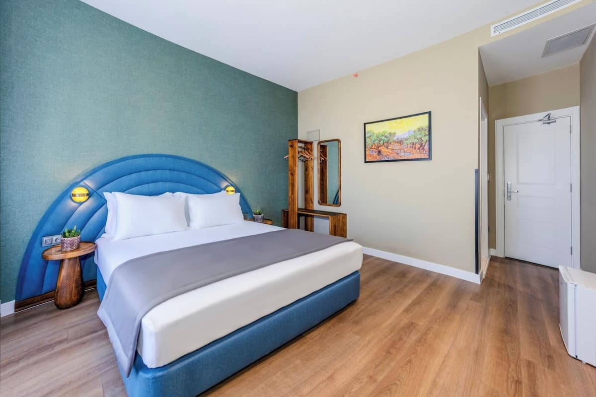 A bedroom with a large bed, a headboard, a bedside table with a lamp, a mirror, and a door.